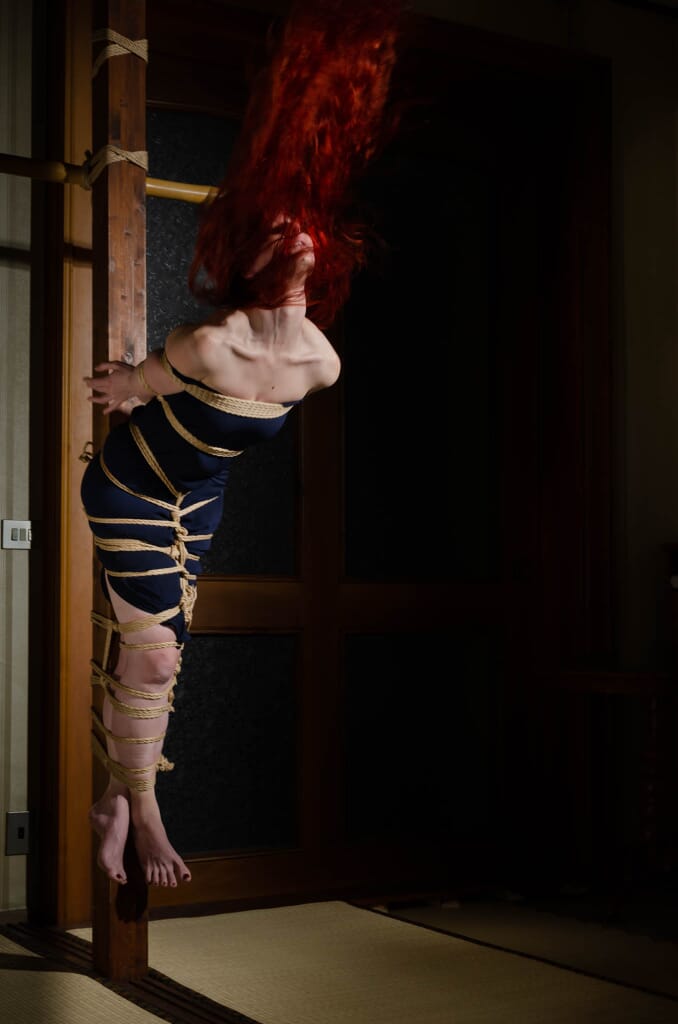 Marta si tende fuori asse dall’hashira mentre i capelli rossi si sollevano nel movimento, nella progressione di kinbaku