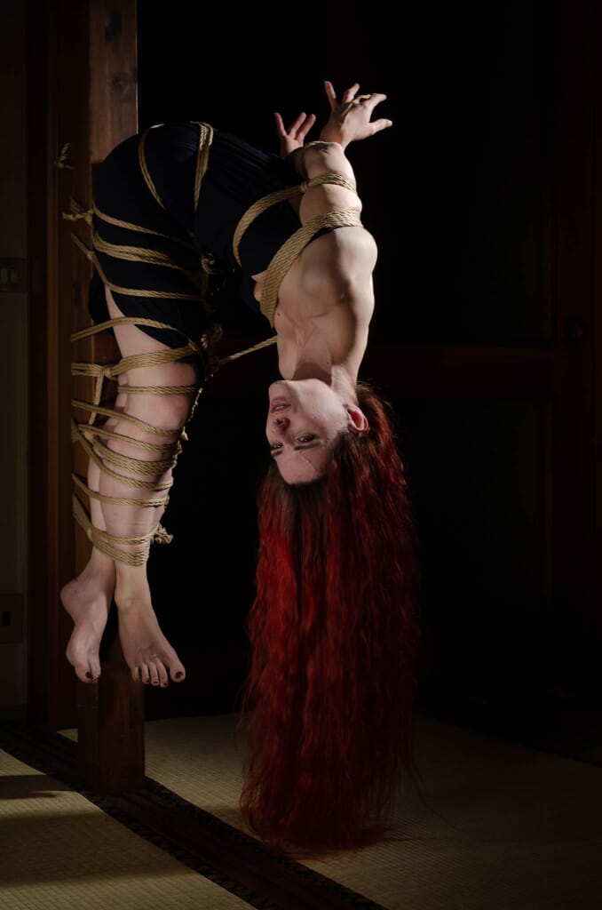 Marta sospesa a testa in giù con il corpo curvo e i lunghi capelli distesi, nel contesto del kinbaku