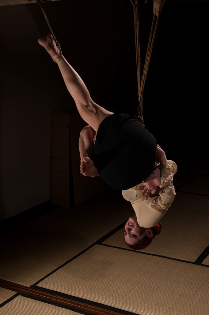 Marta guarda la camera durante una sospensione capovolta nel vestito sobrio, nella progressione di kinbaku