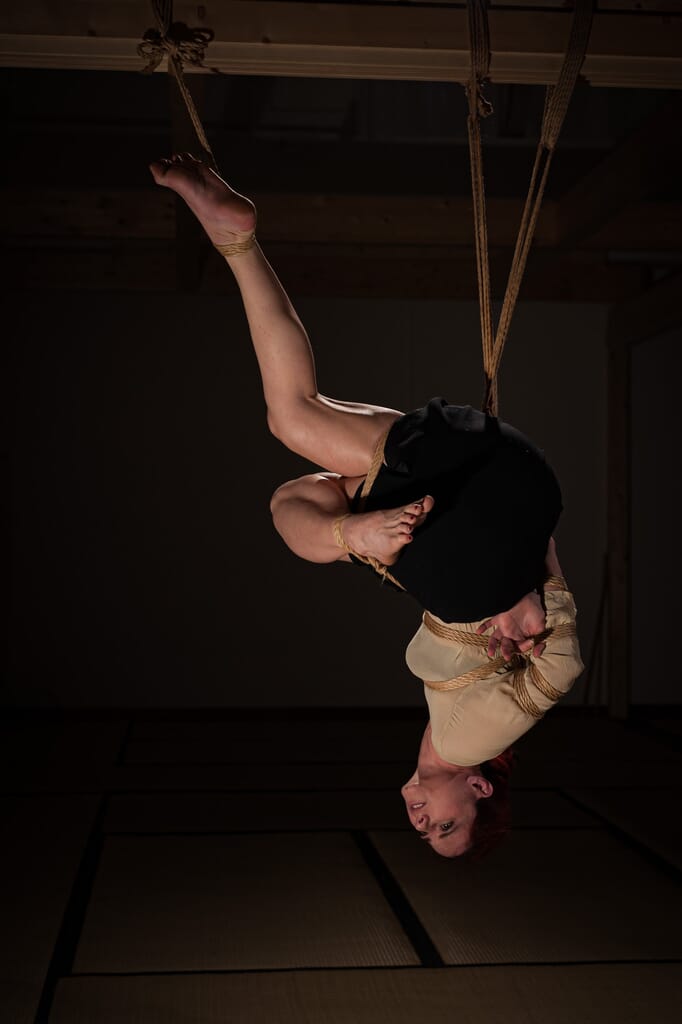 Marta capovolta in sospensione con una gamba distesa verso l’alto, nel contesto del kinbaku