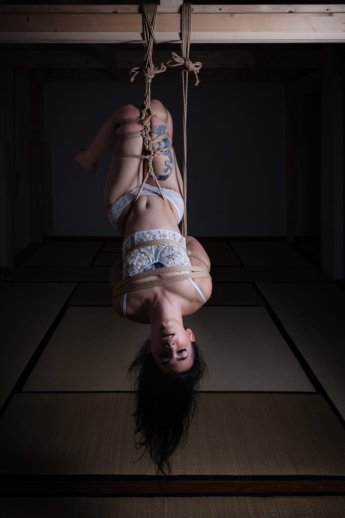 Camelia nella posa legata, con luce radente sul corpo e ombre profonde sullo sfondo, in una sessione di shibari