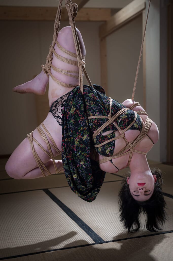 Elettris abbandonata nella sospensione capovolta con il volto sereno, nel contesto del kinbaku