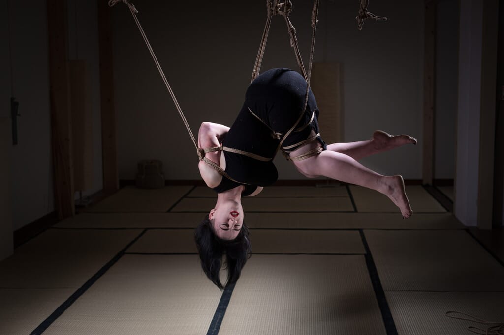 Elettris sospesa a testa in giù in una posizione raccolta, con le corde sul busto e sulle gambe, nel contesto del kinbaku