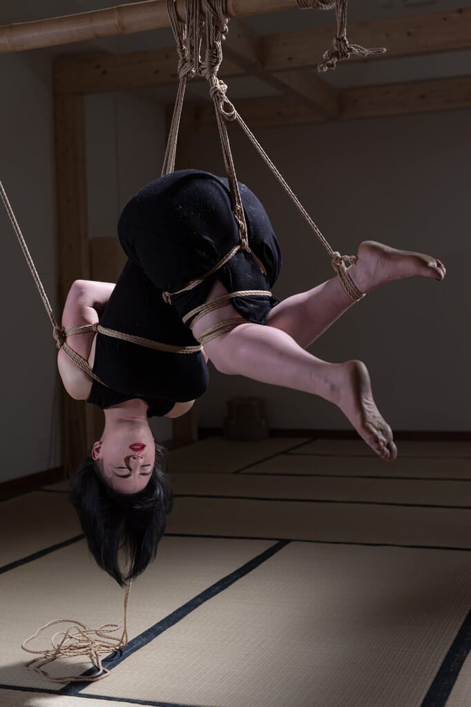 Vista laterale di Elettris nella sospensione capovolta, con una gamba distesa verso l’esterno, nella progressione di kinbaku
