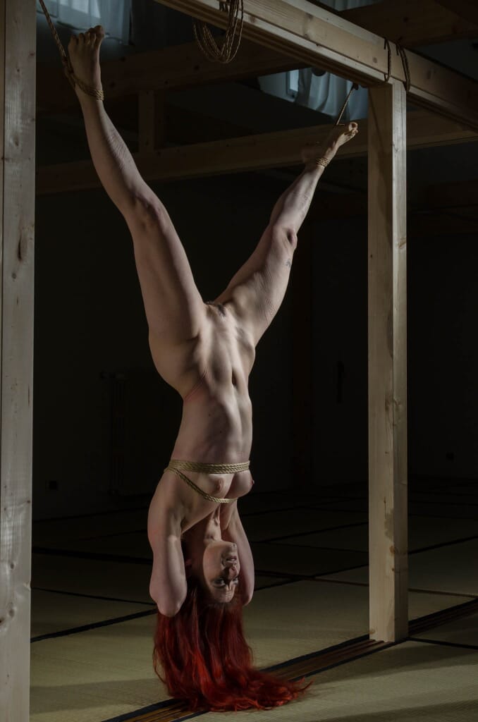 Marta Tenshiko sollevata per le caviglie accanto all'hashira all'inizio della sospensione, nel contesto del kinbaku