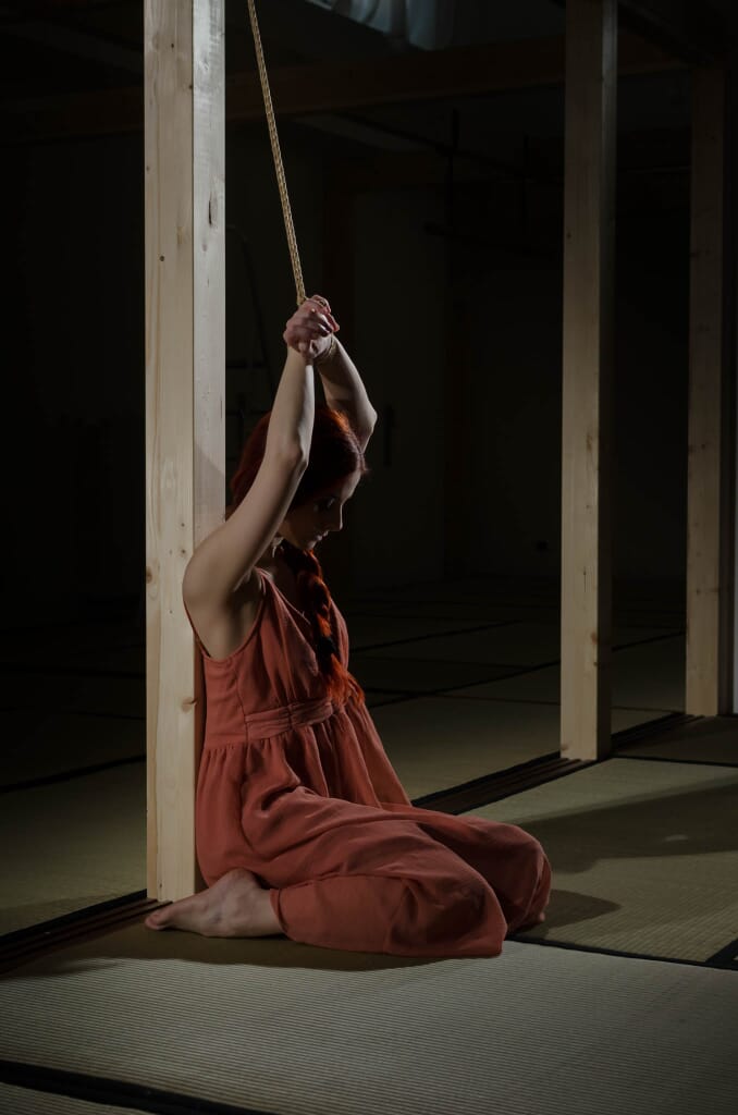 Figura intera di Marta Tenshiko legata all'hashira, con le curve del corpo contro la linea verticale del palo