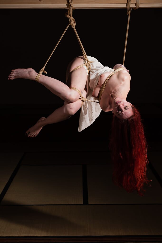 Marta Tenshiko suspended sideways with her torso arched and one arm reaching toward the tatami