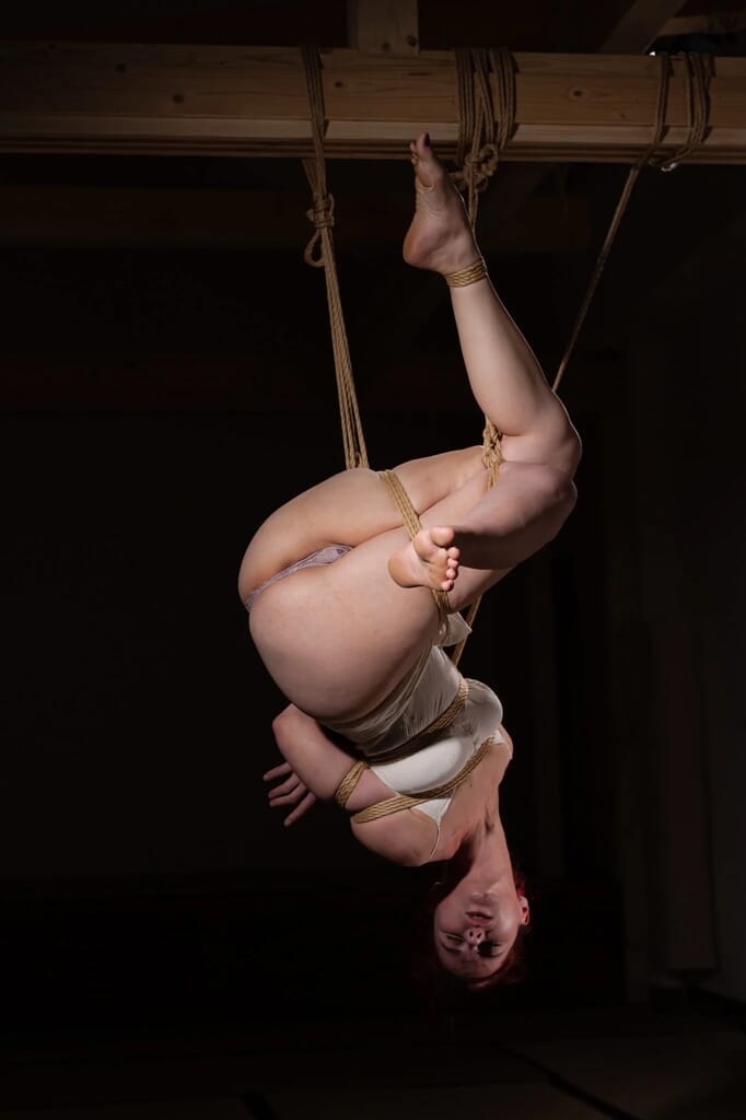 Marta Tenshiko folded into an inverted suspension, with her hips raised and knees bent, within a kinbaku session