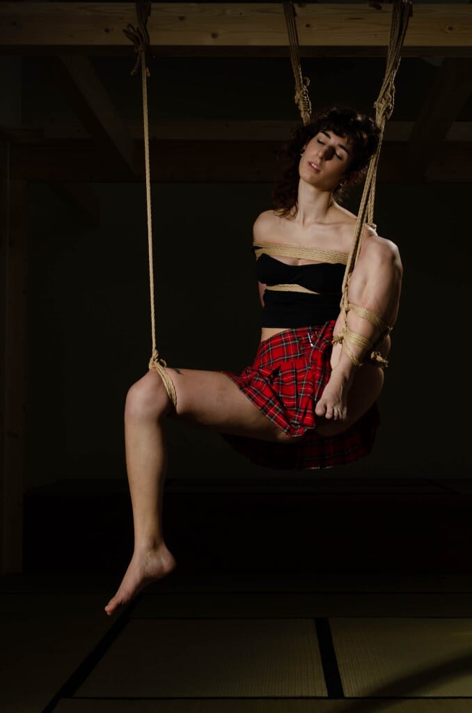d.i.f.y.s. sospesa con il busto legato e una gamba sollevata, illuminata contro il fondo scuro, nella progressione di kinbaku
