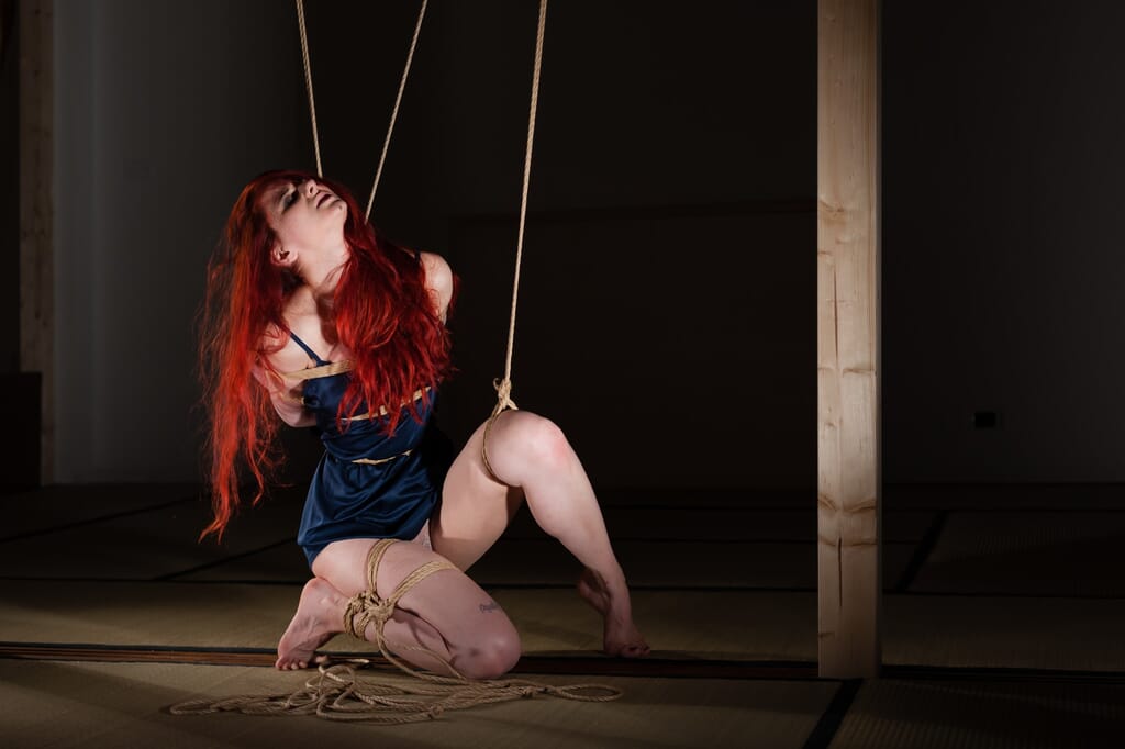 Marta Tenshiko risale in ginocchio seguendo una linea di corda e guarda verso l'alto, nello studio fotografico di shibari