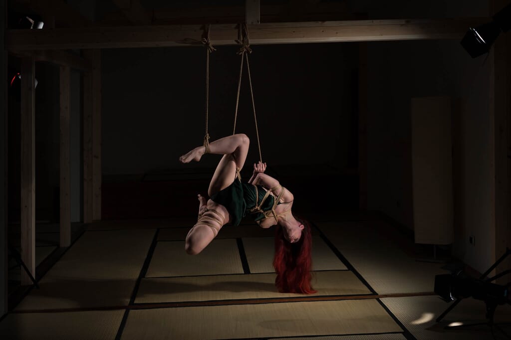 Marta Tenshiko ruota nella sospensione capovolta, incorniciata dalle linee verticali delle corde, nella progressione di kinbaku