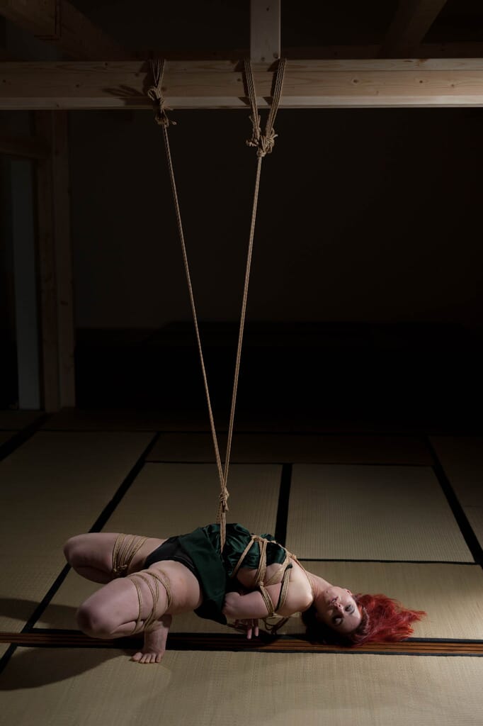 Vista intera di Marta Tenshiko in appoggio su un piede, con l'altra gamba legata e sollevata, nella progressione di kinbaku