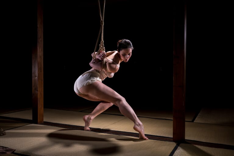 Marta Tenshiko avanza in una sospensione shibari bassa, con una gamba tesa e l’ombra sul tatami